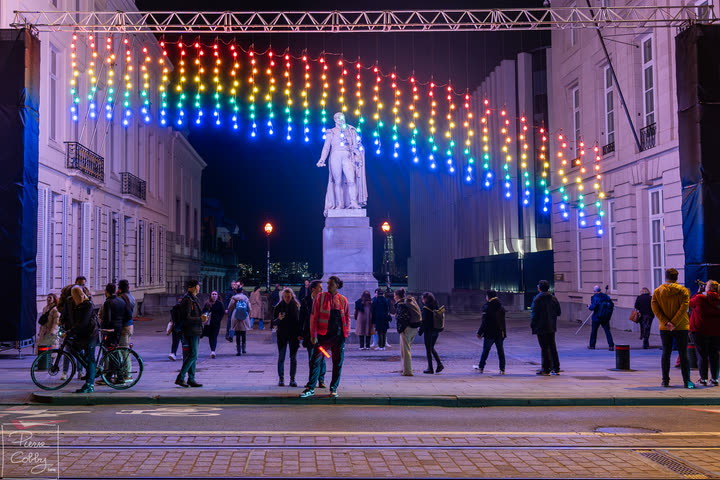 Rainbow light installation over a statue and a crowd at night, Brussels